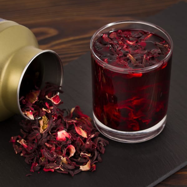 A glass of red hibiscus tea next to a gold tin spilling dried hibiscus petals.