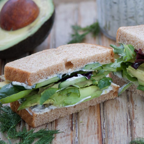 Two avocado and lettuce sandwiches on whole wheat bread with a creamy spread, on a wooden surface.